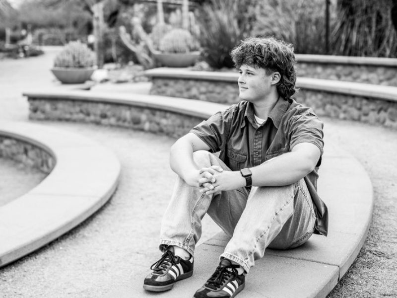 Black and white senior portrait of a high school boy sitting on a stone ledge in a garden, photographed by Andria May Photography in Gilbert, Arizona