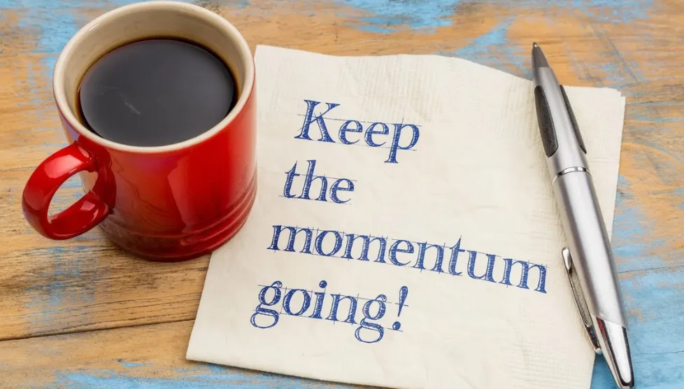 Red coffee cup on wooden desk beside a napkin with the words ‘Keep the momentum going,’ illustrating the blog ‘Why a 90‑Day Action Plan Changes Everything’ about turning retreat insights into lasting leadership improvements.