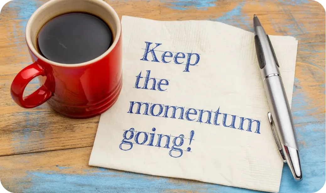 Red coffee cup on wooden desk beside a napkin with the words ‘Keep the momentum going,’ illustrating the blog ‘Why a 90‑Day Action Plan Changes Everything’ about turning retreat insights into lasting leadership improvements.