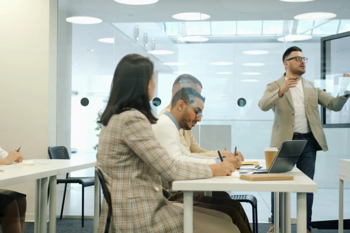 Modern office meeting room with a presenter speaking to a small group seated at white tables, taking notes and listening attentively in a bright, collaborative setting.