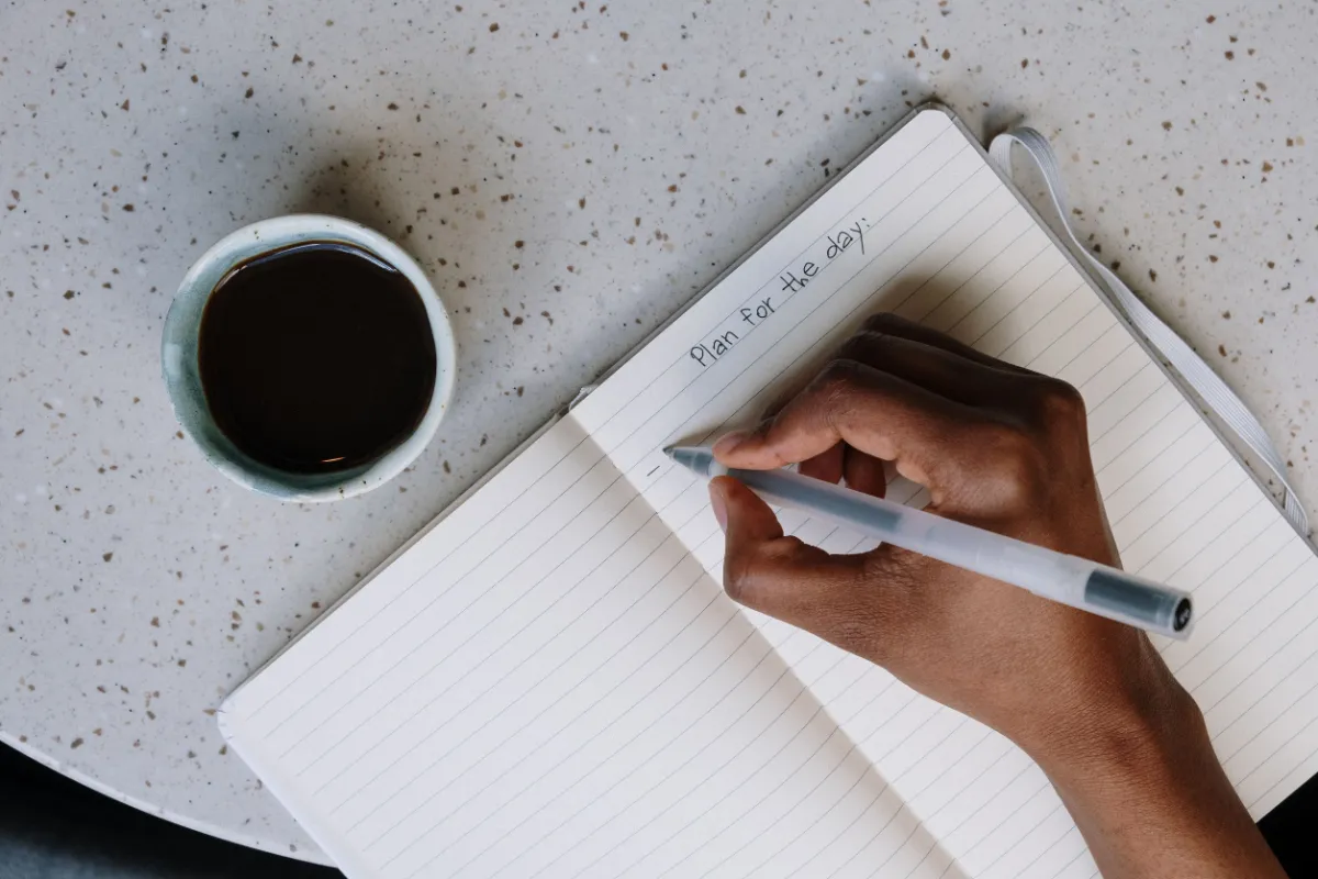 Hand writing a daily plan in a notebook beside coffee, symbolizing the programme’s focus on turning insight into five practical commitments and a structured 90‑day plan.