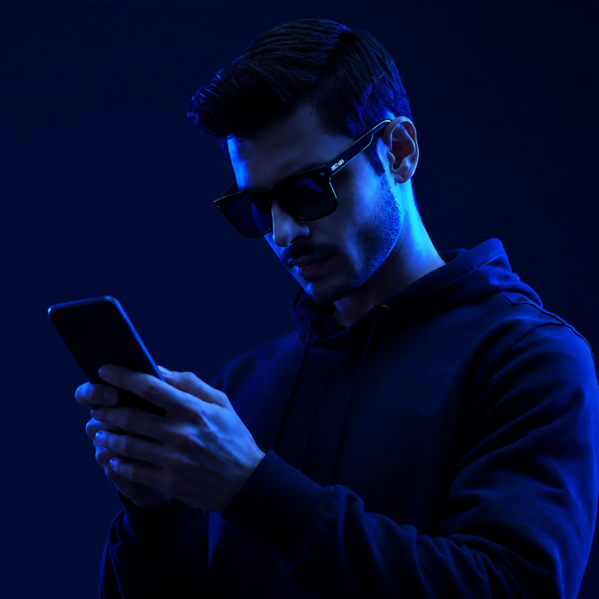 A man in a dark hoodie wearing sunglasses looks down at his smartphone, illuminated by purple and blue neon lighting.
