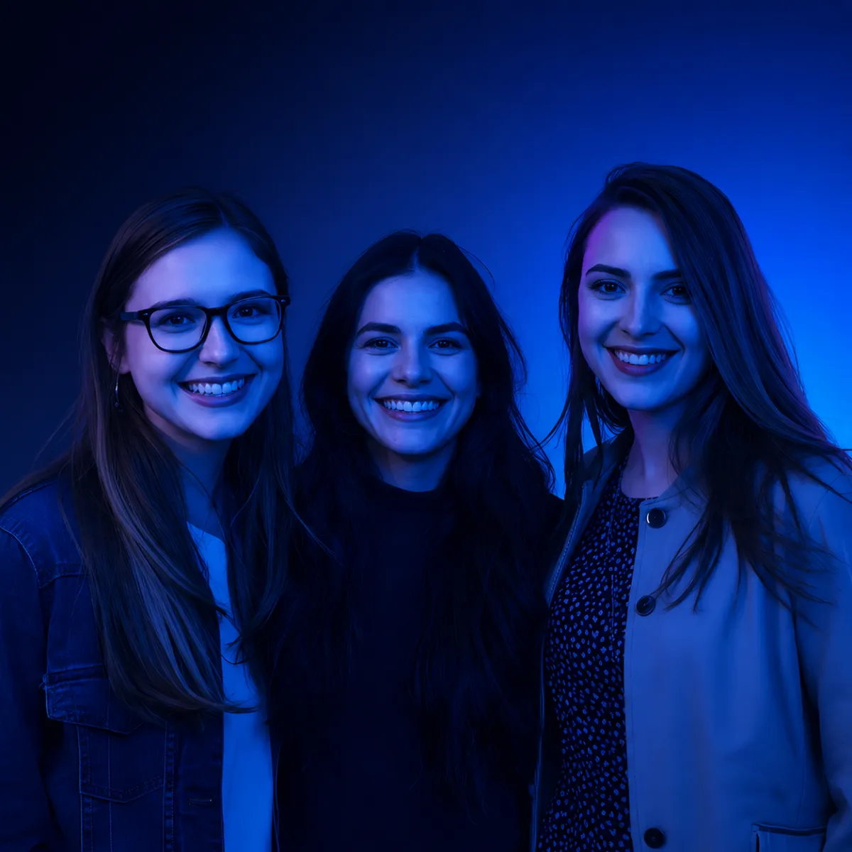 Three young women standing close together and smiling, illuminated by soft blue and purple lighting against a plain background.