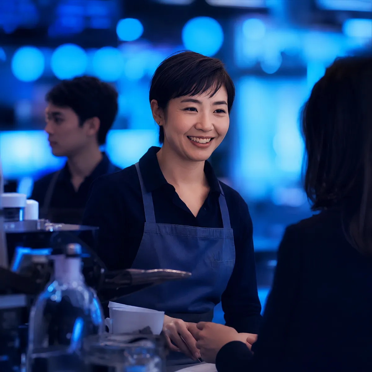 A barista wearing an apron smiling while interacting with a customer across the counter, shown in a purple-tinted photographic style.
