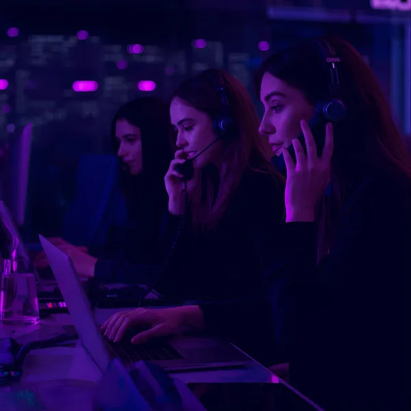 Three women wearing headsets working at computers in a dimly lit call center with purple ambient lighting.