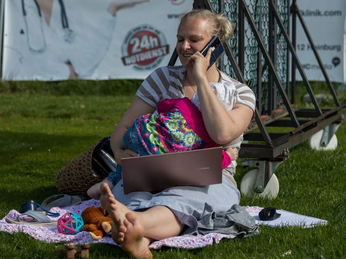 Mama mit Kind im Arm auf Fußballplatz sitzend, Laptop auf dem Schoß, Kind liegend im Arm und telefonierendp
