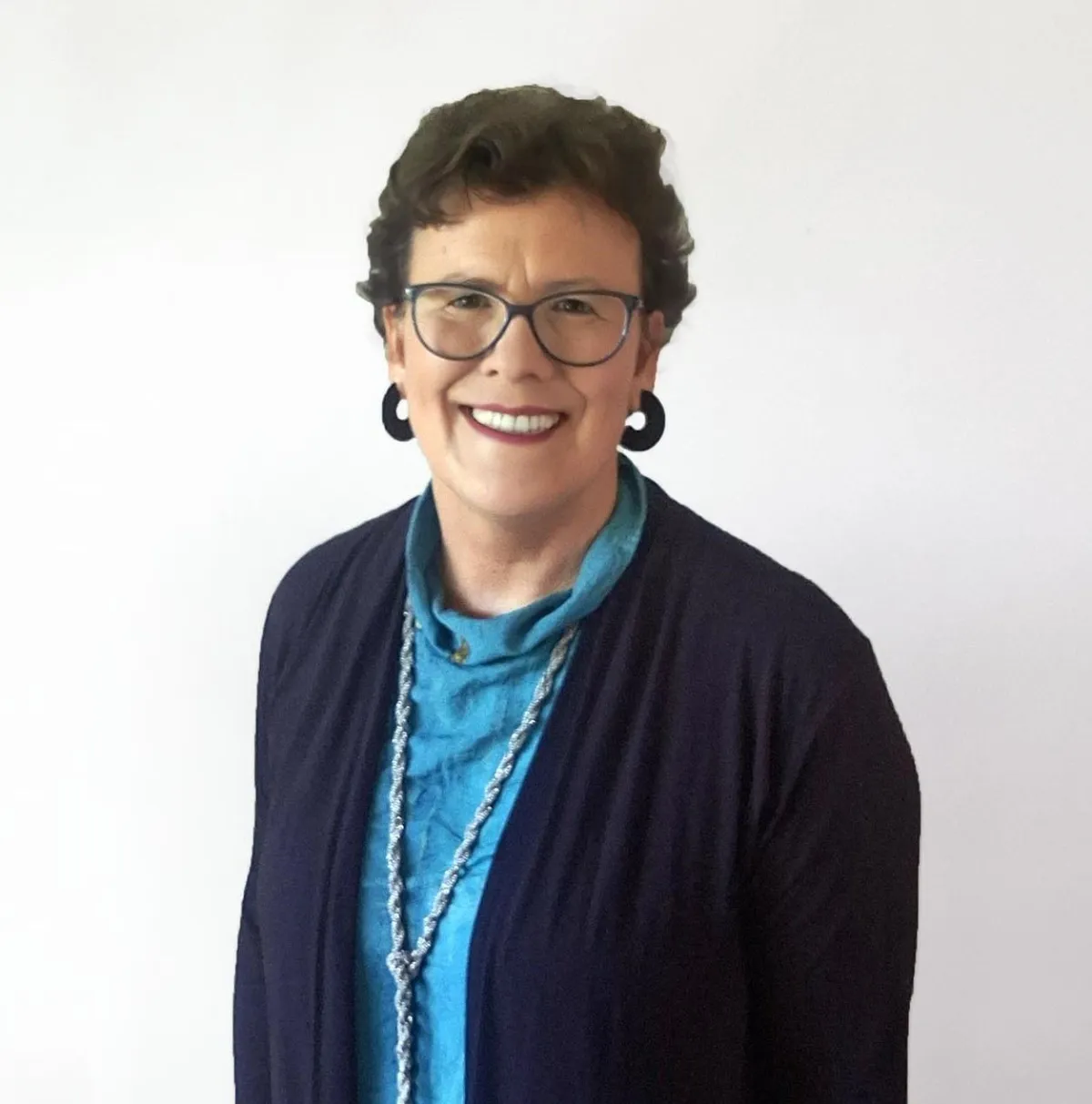 Portrait of Jean Kropper, a smiling woman with short curly hair, wearing glasses, a blue top, a navy cardigan, and a long necklace, standing against a plain white background.