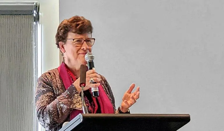 Jean Kropper speaking at the 'Women with Altitude Conference' in Penrith, NSW, Australia, wearing a red scarf and glasses while standing at a podium.