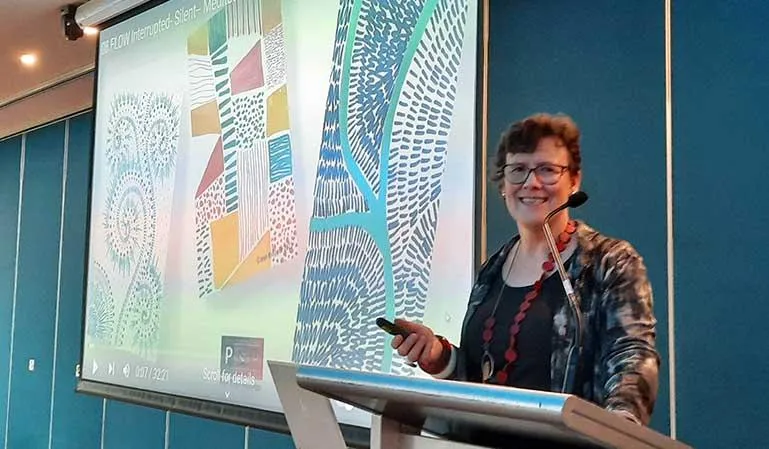Jean Kropper speaking at the 'City of Whittlesea' event in Victoria, Australia, presenting a meditative drawing workshop to council employees, with a large screen displaying abstract artwork behind her.