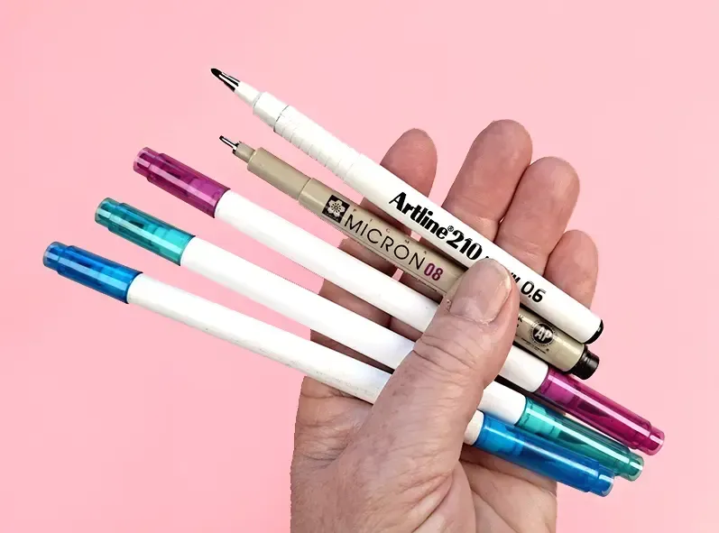 A close-up of a hand holding various drawing pens, including fine liners and colored markers, against a pastel pink background, symbolizing the tools used in Meditative Drawing.