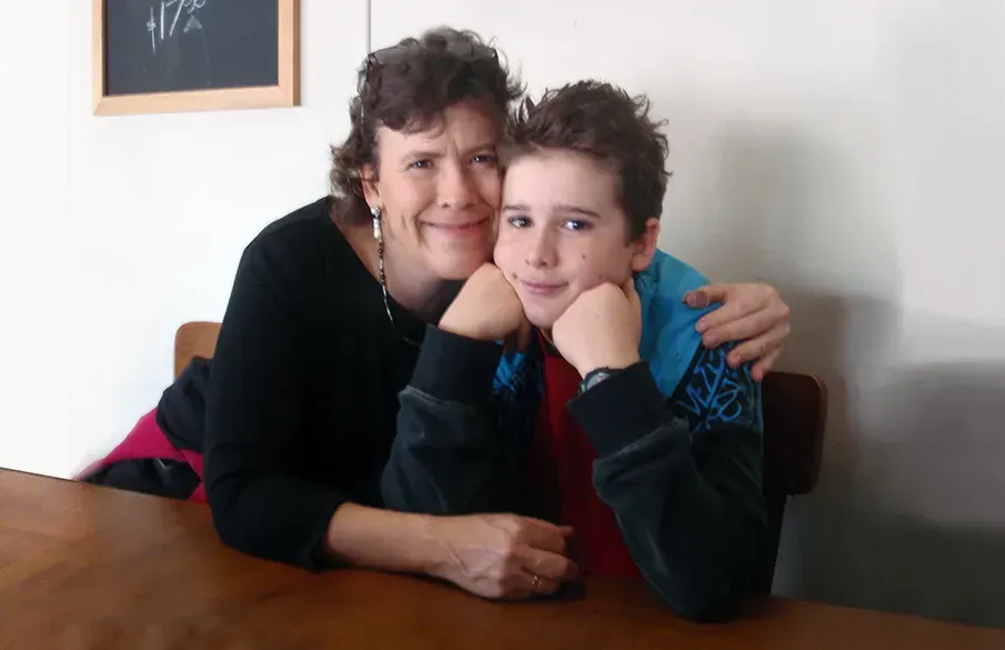 Jean Kropper sitting at a wooden table with her son in 2014, both smiling warmly and leaning on their hands, with Jean’s arm wrapped around him in an affectionate pose