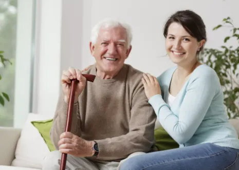 Smiling elderly man holding a cane, seated beside a young woman in a cozy home setting, reflecting compassionate healthcare and family support.