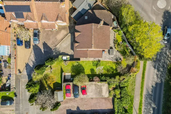 An ariel birds eye view of a property. You can see the roof as well as neighbouring roofs. There are driveways, a road, and parked vehicles. The lawn, trees, all in green from Spring and Summer.