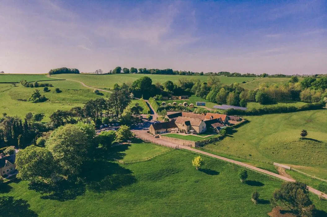 Aerial photograph of a Cotswold stone farm set amongst rolling green fields with trees and haybales, taken on a sunny day with blue sky and trees on the horizon