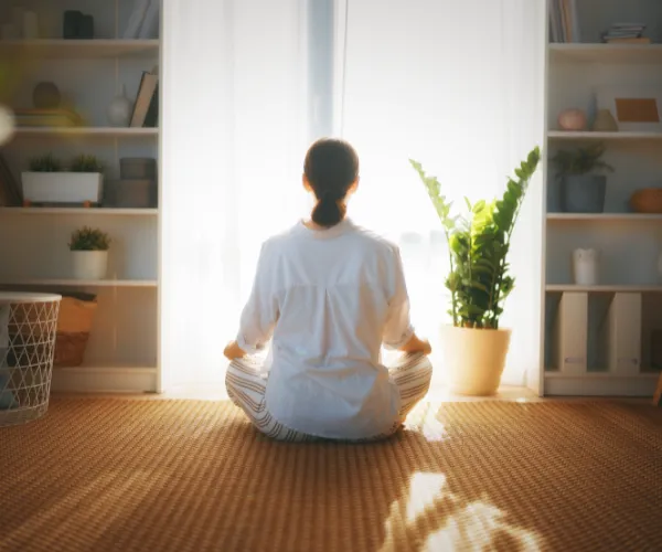 A woman sitting in quiet, mindful meditation in front of a window illuminated by soft morning light, practicing somatic calming exercises.