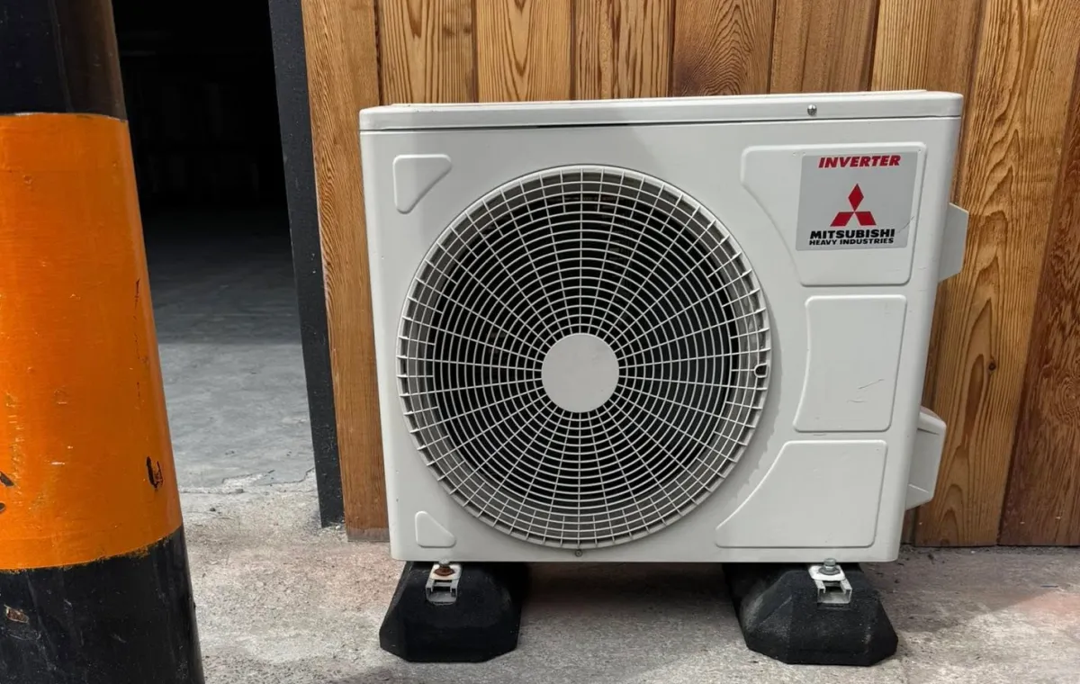 Outdoor air conditioning unit against a wooden wall, next to a black and orange pole. The unit is a Mitsubishi Inverter model on concrete blocks.
