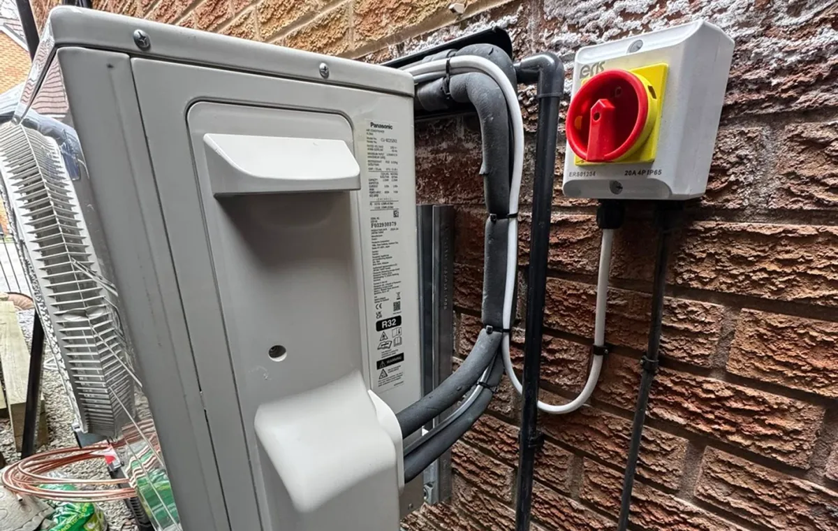 Outdoor air conditioning unit attached to a brick wall. Nearby are cables and an electrical box with a red switch. The scene appears technical and industrial.