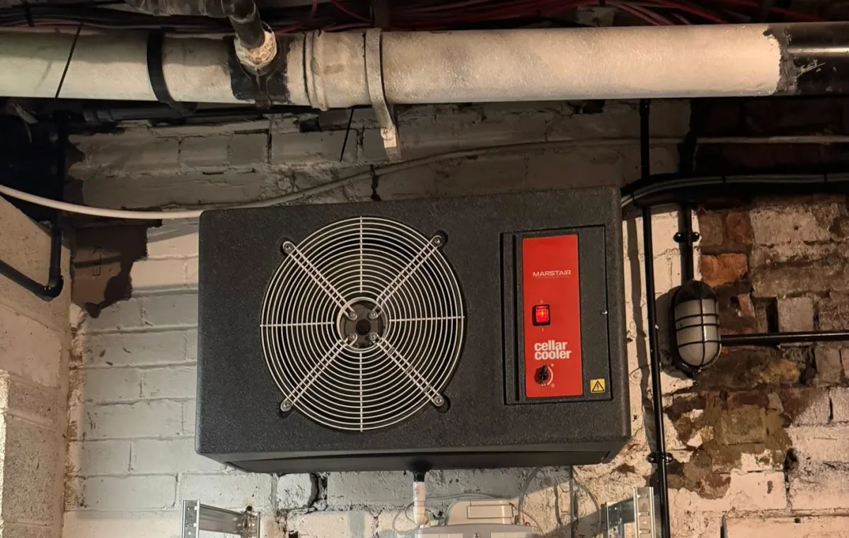 Industrial ventilation unit mounted on a brick wall in a cellar, featuring a large fan and a red control panel. Exposed pipes and cables surround it.