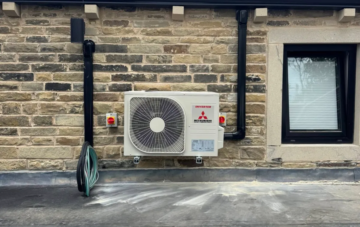 Outdoor air conditioning unit mounted on a stone brick wall beside a window. A coiled green hose is placed on the left, and the scene conveys a functional, neat appearance.