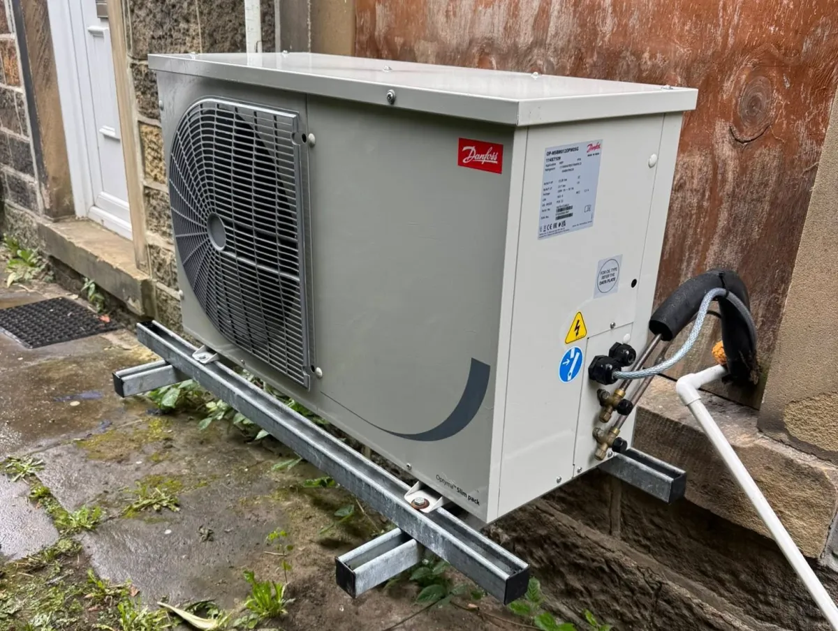 Outdoor HVAC unit mounted on metal brackets against a stone wall, with visible pipes and wiring. The ground below is damp and overgrown with weeds.