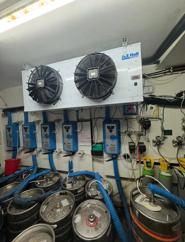 A room filled with beer kegs connected to cooling units on the ceiling. Blue hoses and wires are visible, creating an industrial setup. Lighting is fluorescent.
