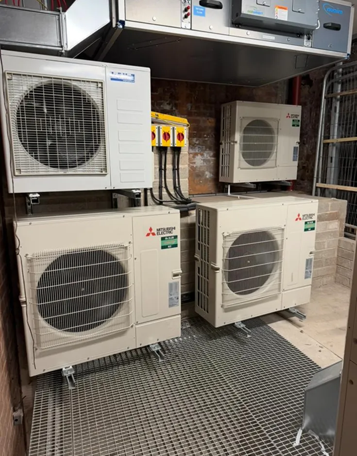 Indoor utility room with four white HVAC units mounted on a metal grid floor. Visible cables and controls suggest an industrial setting.