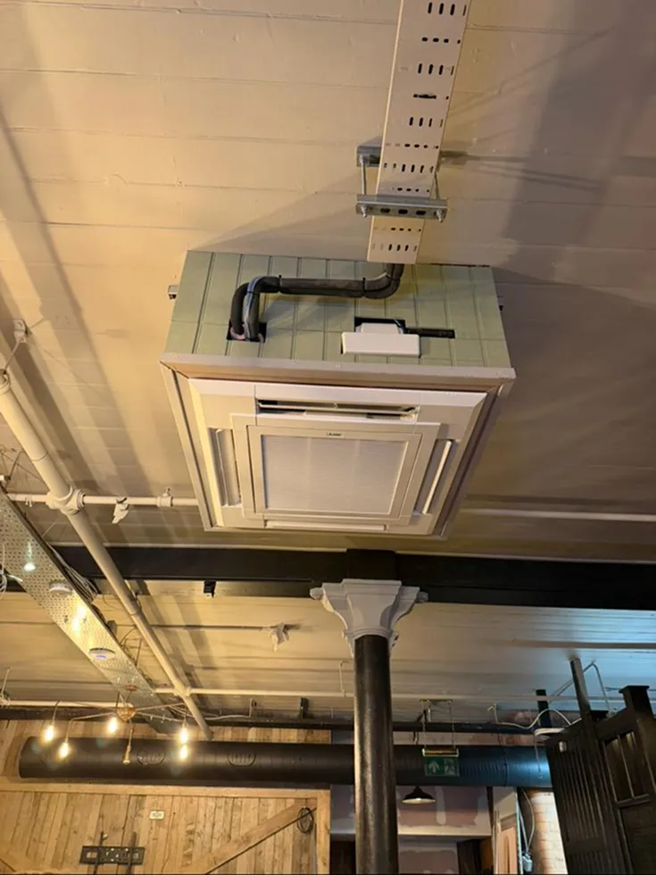 Ceiling view of an inverted room with a mounted air conditioner unit and exposed pipes. Wooden panels and industrial beams add a rustic feel.