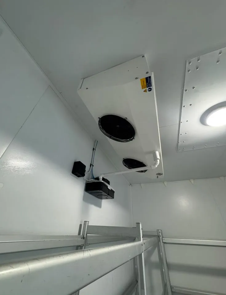 A refrigerated room interior with metal shelves, a ceiling vent with two fans, and a small light. The space is sterile and industrial.