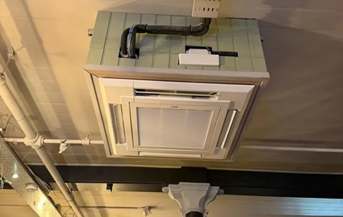 Ceiling-mounted air conditioning unit with a vent surrounded by light green tiles. Exposed pipes and beams create an industrial feel.