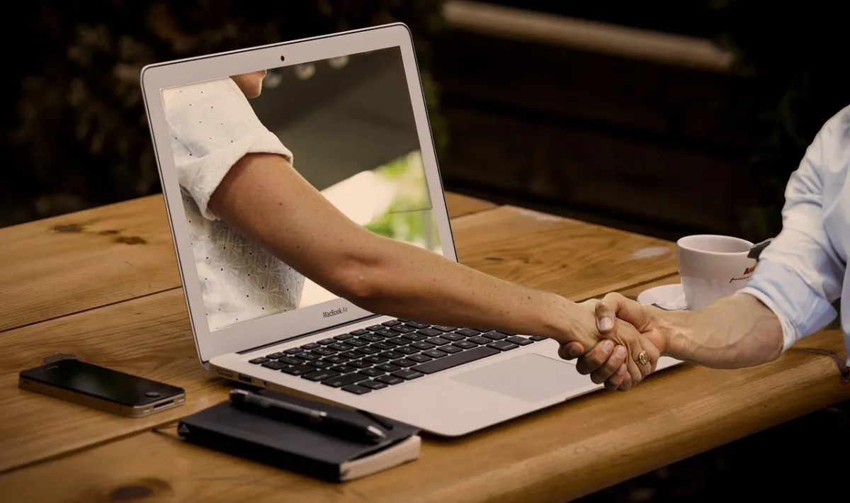Team members joining hands over a shared wooden office desk with laptops, coffee, notebooks and a small plant—symbolizing workplace collaboration, unity, and team spirit.