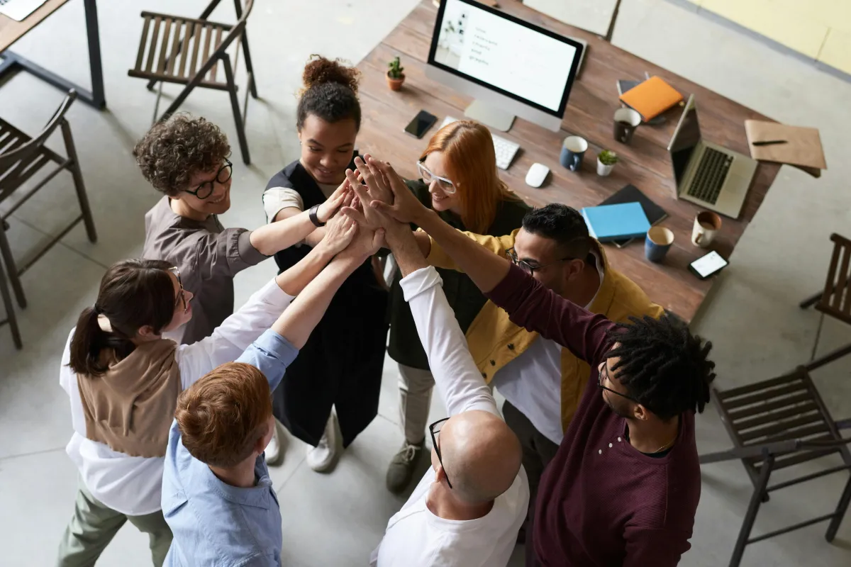 Team members joining hands over a shared wooden office desk with laptops, coffee, notebooks and a small plant—symbolizing workplace collaboration, unity, and team spirit.