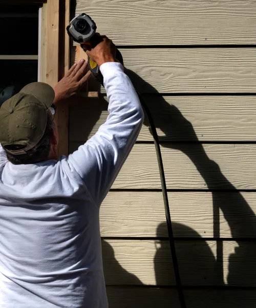 A worker wearing a cap and a white long-sleeved shirt is using a power tool to cut a piece of siding or trim on the exterior of a building.