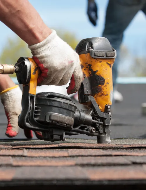 A close-up of a roofer's gloved hands using a nail gun to fasten shingles to a roof.