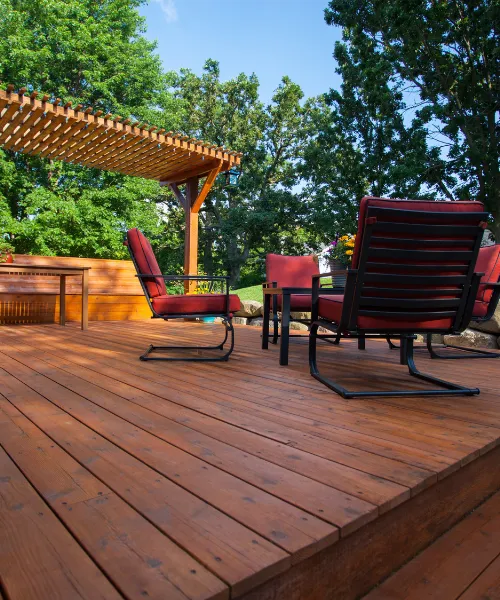 low-angle view of a wooden deck with a slatted pergola, surrounded by trees. The deck has a patio set with metal chairs and red cushions.