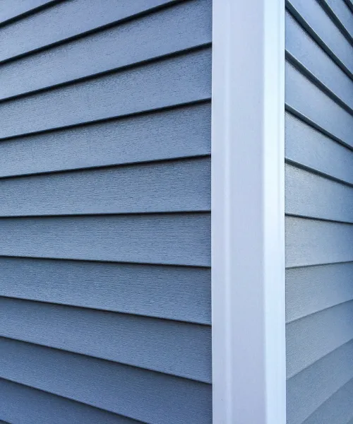 A close-up of the corner of a house, showing textured blue horizontal siding and a smooth white vertical trim piece.