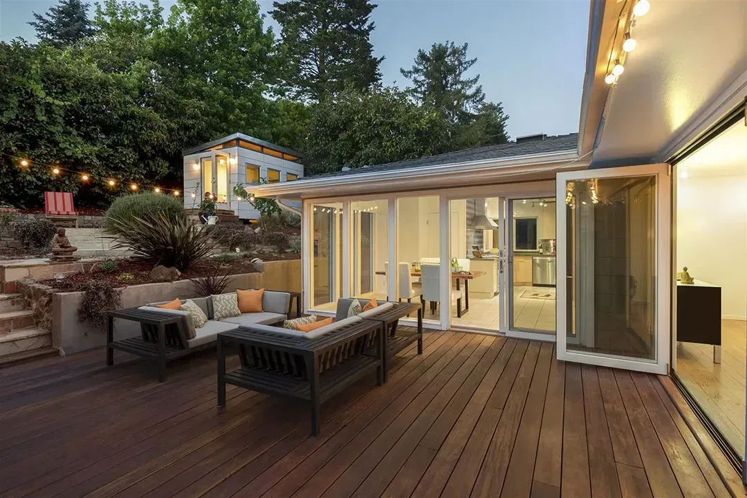 modern wooden deck with a large sectional couch and an outdoor coffee table. The deck is attached to a house with large sliding glass doors, which are open to a dining area and kitchen. 