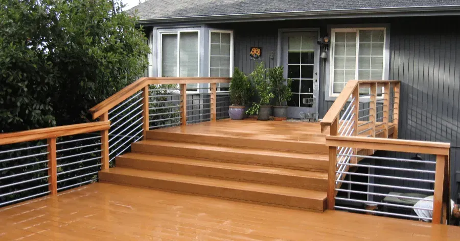A large wooden deck and staircase with a light brown finish. The deck railings are made of wood with horizontal metal balusters. The deck is attached to a gray house with a glass door and multiple windows. 