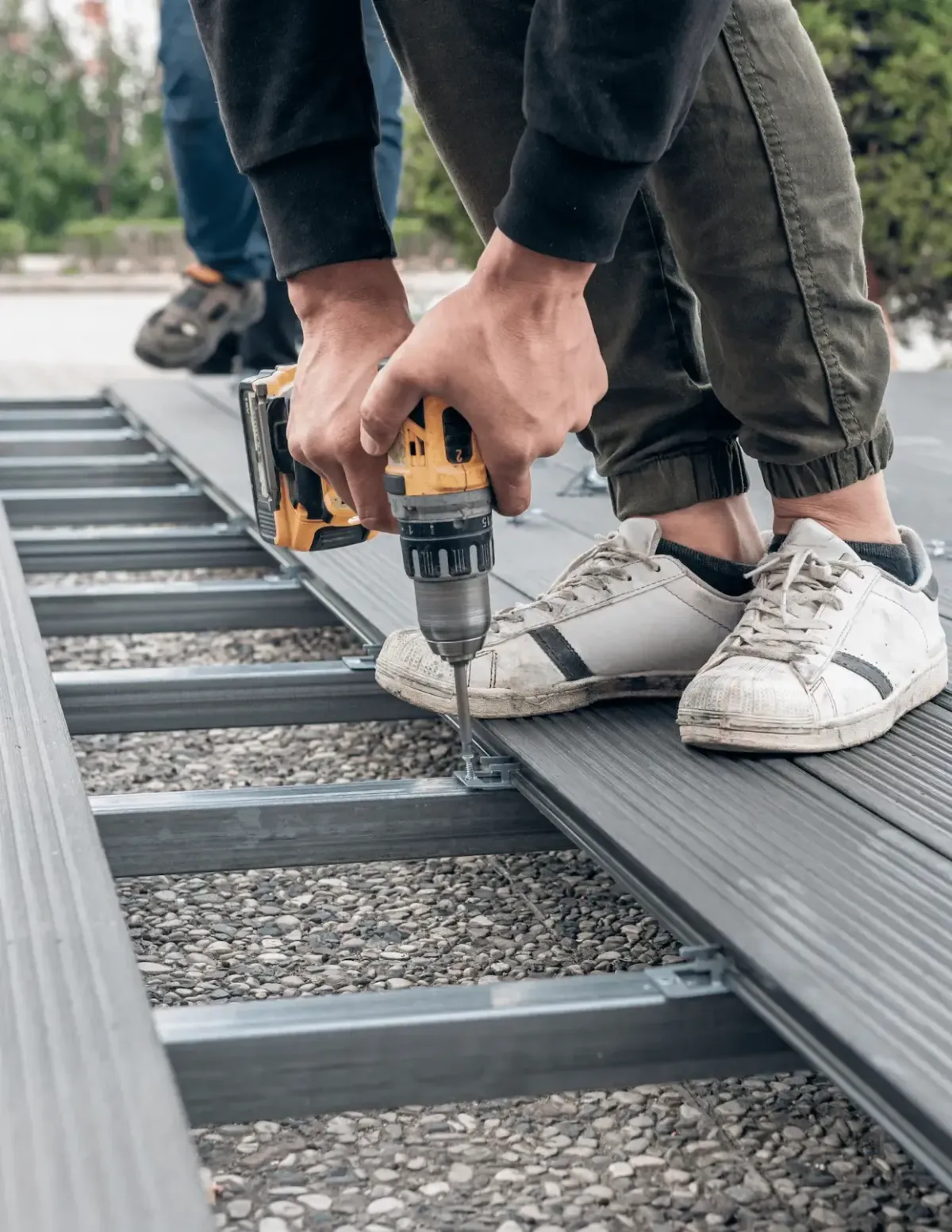 A close-up shot of a person using a yellow and black power drill to install gray composite decking onto a metal subframe. 