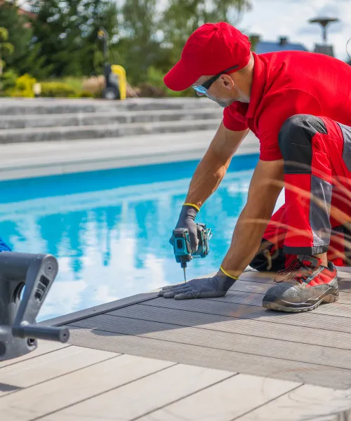 A person in a red shirt, red pants, and red cap is using a power drill to install composite decks along the edge of a swimming pool.