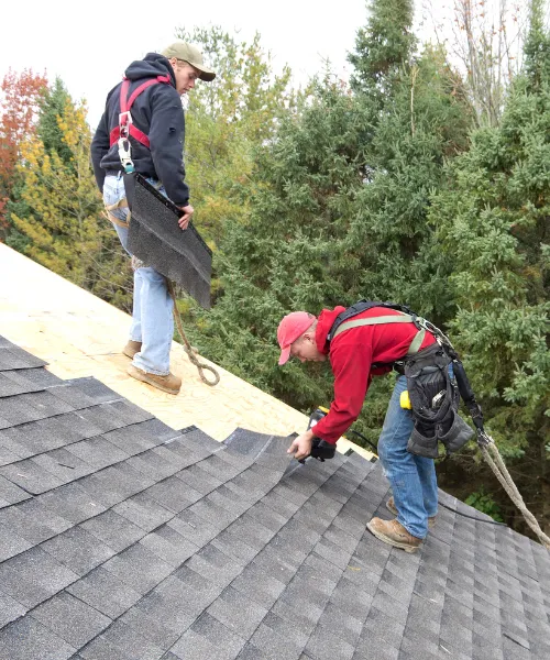 Two roofers, wearing safety harnesses and tool belts, are installing shingles on the steep roof of a house, surrounded by trees.