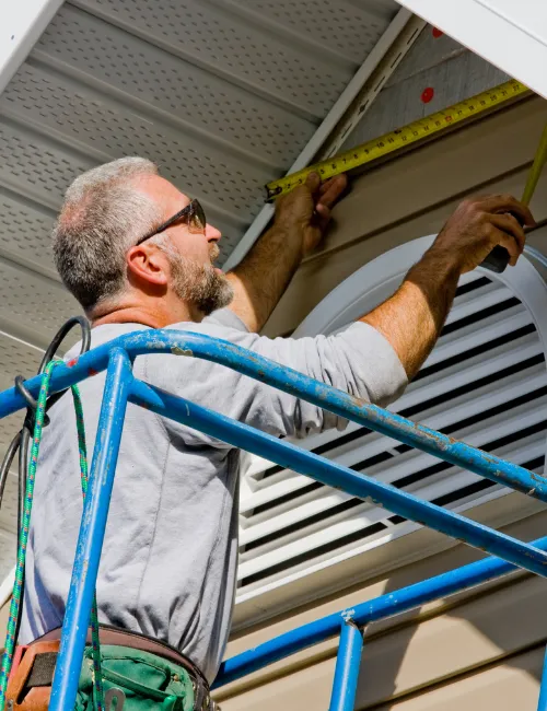 A male worker on a scaffold is using a tape measure to measure the dimensions of a building's soffit and siding. 
