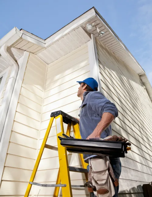 A siding contractor on a yellow ladder is looking up to inspect the gutters and siding on the corner of a house.