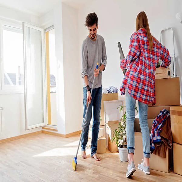 A young man sweeping a hardwood floor with a broom while a woman stands near cardboard moving boxes holding a roll of packing material in a bright room.