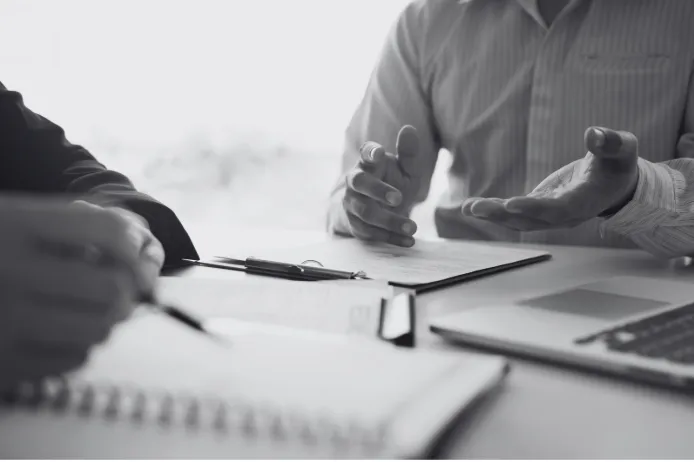Two colleagues discussing a workplace document during a meeting, with notebooks and a laptop on the table.