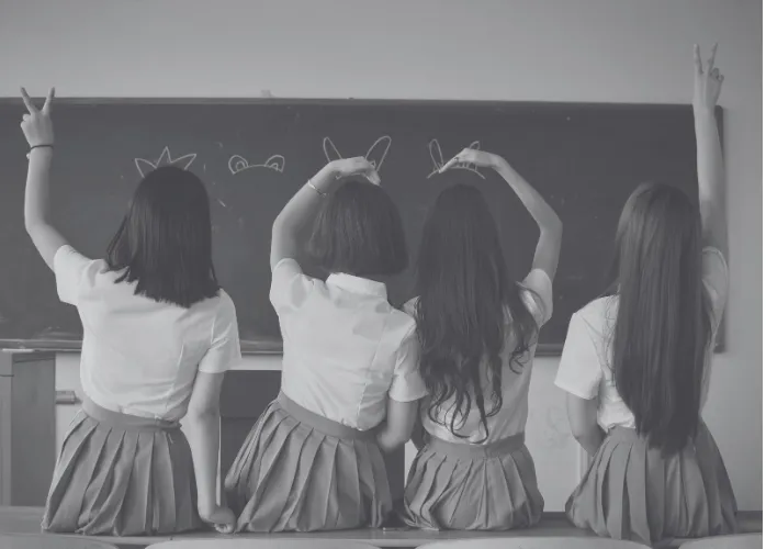 Four schoolgirls sitting with their backs to the camera and making playful gestures in front of a classroom chalkboard.