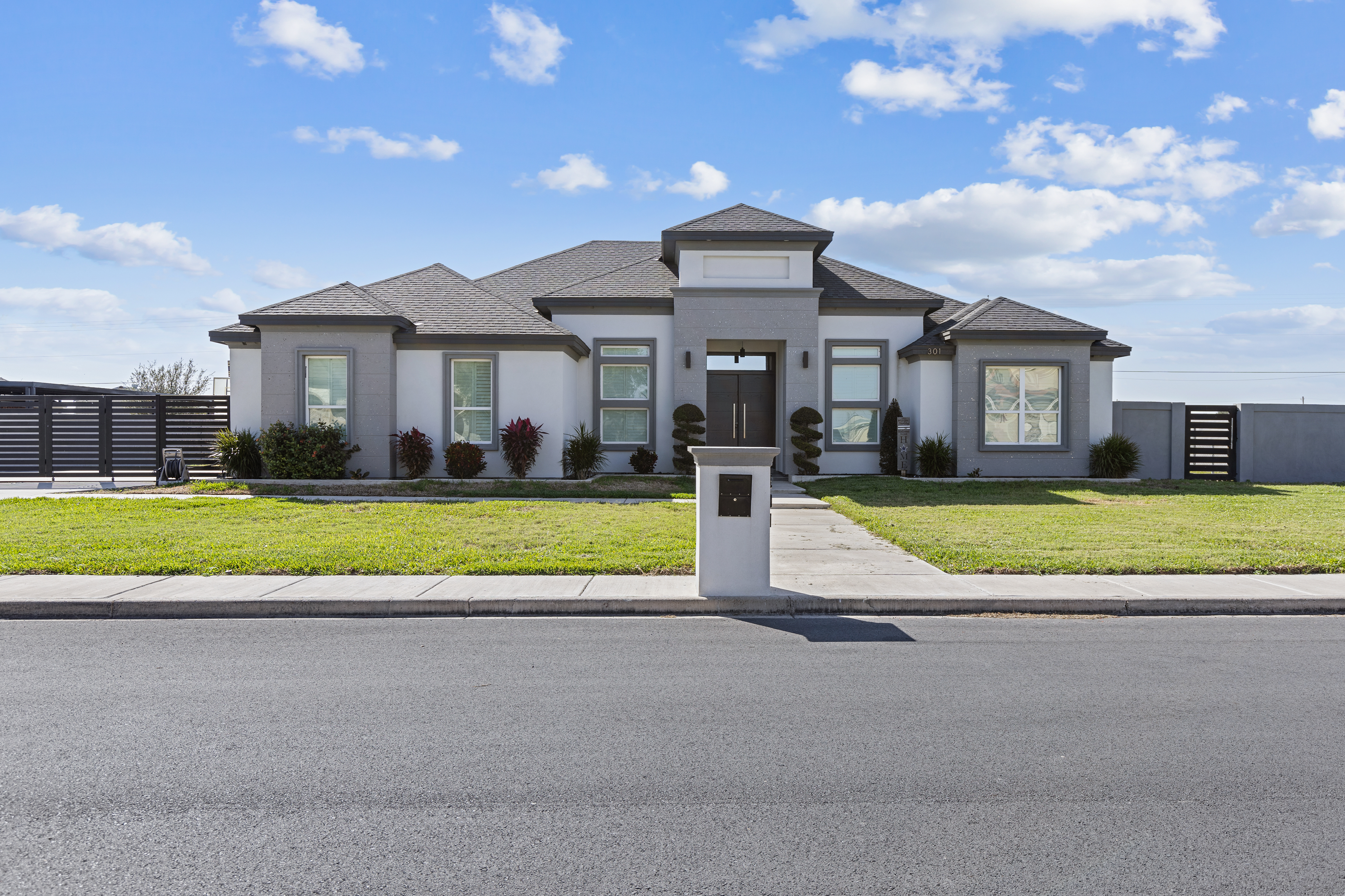 Modern gray custom home exterior with white stucco, dark trim, front lawn, and gated driveway in South Texas.