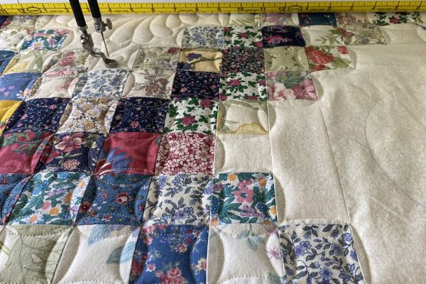 Nærbilde av quilt, firkanter i blomstrete stoff med quiltede buer, lyst uquiltet felt til høyre. Foten på longarmmaskinen sees såvidt oppe i venstre hjørne