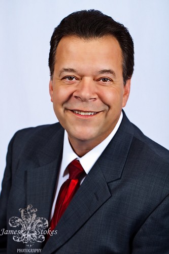 Don Everhard, a confident middle-aged business speaker in a tailored suit, stands on a stage with a warm spotlight, gesturing as he addresses an engaged audience. The background is softly blurred, emphasizing his approachable demeanor and professional presence.