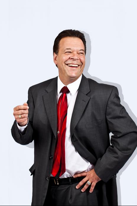 Don Everhard, a confident middle-aged business speaker in a tailored suit, stands on a stage with a warm spotlight, gesturing as he addresses an engaged audience. The background is softly blurred, emphasizing his approachable demeanor and professional presence.