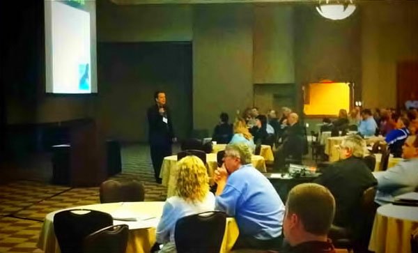 Don Everhard, a confident middle-aged business speaker in a tailored suit, stands on a stage with a warm spotlight, gesturing as he addresses an engaged audience. The background is softly blurred, emphasizing his approachable demeanor and professional presence.